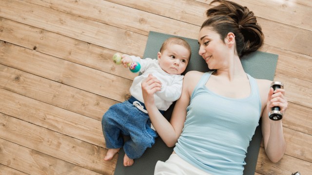 a mom in workout gear lays on a green yoga mat with a weight, newborn next to her, fourth trimester activities