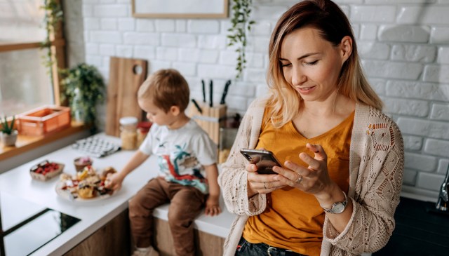 a mom on her phone looking at a family organizer app while her toddler is in the background looking at a tray of desserts